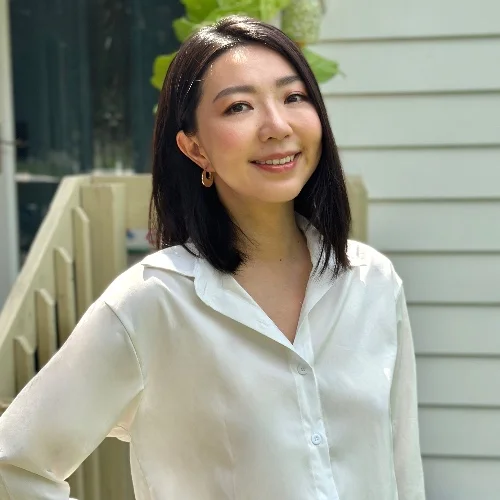 a photo of a smiling woman wearing a white button-up shirt standing in front of a light-colored house.